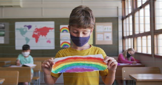 Caucasian child in yellow shirt and mask holding a rainbow drawing in a classroom lifts spirits during COVID-19 pandemic. Diverse students in background engaged in their tasks, showcasing productive classroom environment. Useful for concepts of post-COVID schooling, children's creativity, education adaptations, and multicultural classrooms in the pandemic context.