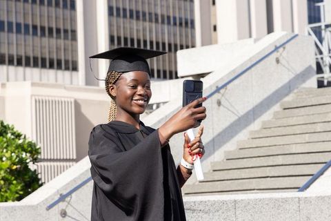 African american graduate taking selfie on urban campus holding diploma and mortarboard