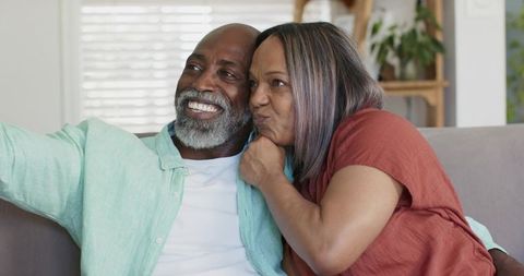 Senior couple taking cozy selfie on living room couch, smiling and cuddling during relaxed moment