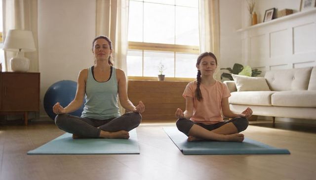Mother and daughter meditating on yoga mats in sunlit living room practicing mindful family bonding