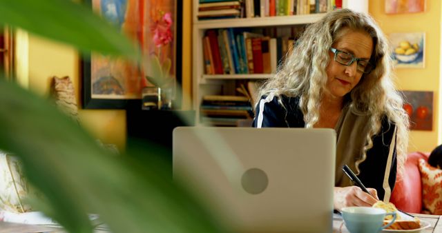 Mature woman with long hair and glasses working at home on laptop, taking notes in a cozy room with various books and artwork in background. Image can be used for articles on remote work, freelancing tips, studying methods, time management, and productivity at home.