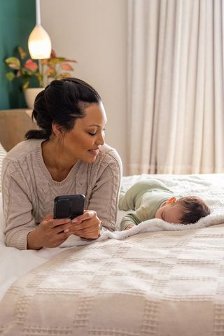 Mother monitoring sleeping baby on bed while using smartphone, tender newborn bonding