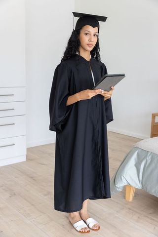Teenage graduate standing in bedroom holding tablet wearing graduation gown and mortarboard