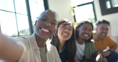 Smiling multicultural friends taking selfie on sunlit living room sofa, candid lifestyle