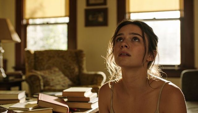 Sunlit young woman gazing upward amid stacks of books in cozy vintage living room, quiet moment