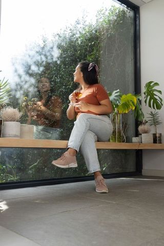 Mid-adult asian woman sitting by sunroom window holding wine glass surrounded by plants