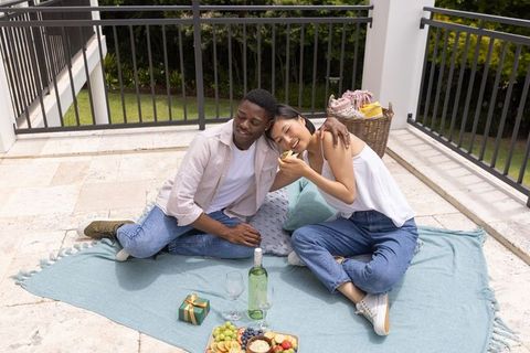 Diverse couple enjoying rooftop picnic sharing fruit and wine on sunny terrace