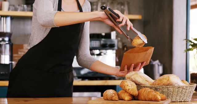 Baker using tongs to place fresh croissants into a paper bag at bakery counter. Background includes bread basket and other baking supplies. Ideal for content related to baking, small businesses, food preparation, and culinary arts. Suitable for advertisements, recipe blogs, or bakery promotions.