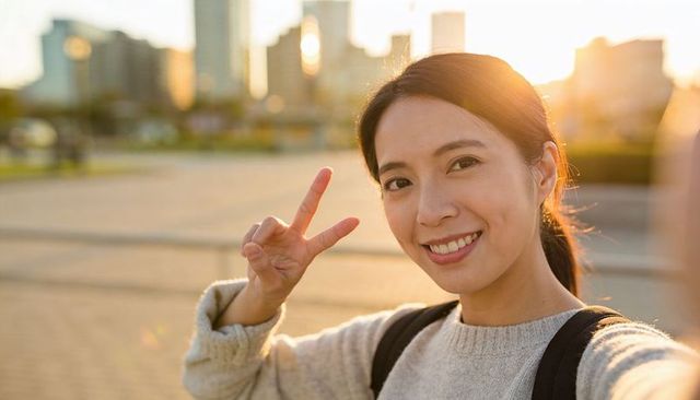 Smiling asian woman taking selfie showing v sign at sunset in urban plaza with backpack