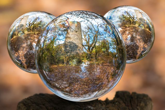 Three glass spheres reflecting an old stone tower and autumn trees, placed on a wooden stump in a forest. Ideal for concepts of reflection, nature, and tranquility. Can be used for nature-themed backgrounds, environmental campaigns, or photography effects tutorials.