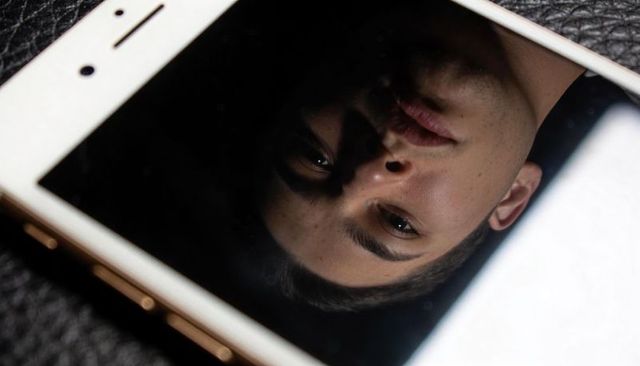 Upside-down teen face reflecting on smartphone screen with dramatic low-light contrast