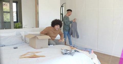 Two men unpacking clothes on bed in sunlit bedroom, organizing wardrobe with hangers and box