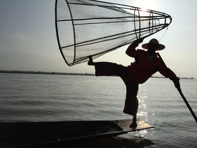 Traditional fisherman balancing on the edge of his boat holding a large fishing net, silhouetted against the sunrise over a serene lake. Ideal for content related to fishing, traditional lifestyles, cultural practices, travel destinations, and outdoor activities.