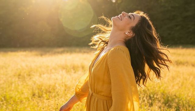 Woman wearing mustard dress embracing golden sunset while smiling in sunlit meadow