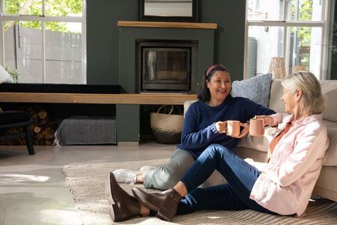 Mature friends sitting on rug sharing coffee by fireplace in cozy modern living room