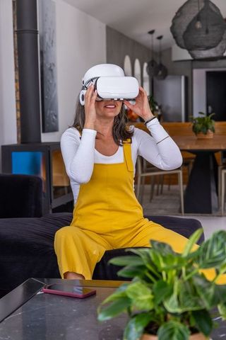 Middle-aged woman exploring virtual reality with vr headset in living room wearing yellow overalls