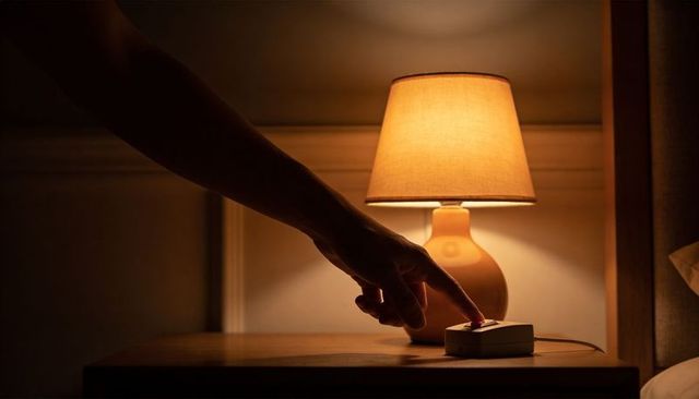 Hand pressing nightstand lamp switch at bedside, turning off warm amber light for sleep ritual