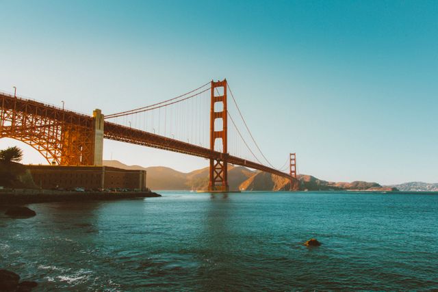 Golden Gate Bridge extending across calm waters during sunset, with a clear sky illuminating the structure. Ideal for use in travel blogs, tourism advertisements, architectural studies, and landscape photography collections.