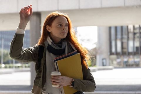Redhead student raising hand on urban campus holding folders and coffee