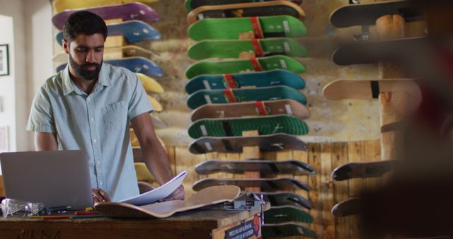 Man reviewing snowboard designs and sketches in a skateboard shop, using a laptop. Ideal for concepts of entrepreneurship, small business, designing sports equipment, or leisure retail operations. Can be used in advertisements, articles on small businesses, or design industry promotions.