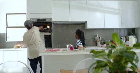 Senior woman reaching into built-in oven while grandchild watches in bright modern kitchen