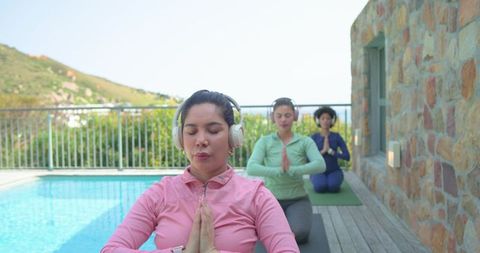 Three women practicing meditation on poolside deck with headphones and yoga mats