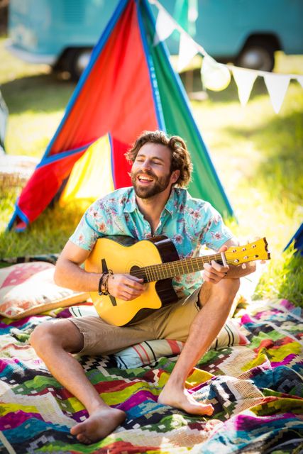 Man enjoying playing guitar at a vibrant campsite on a sunny day. Ideal for use in advertisements, travel brochures, lifestyle blogs, and social media posts promoting outdoor activities, music, and relaxation.