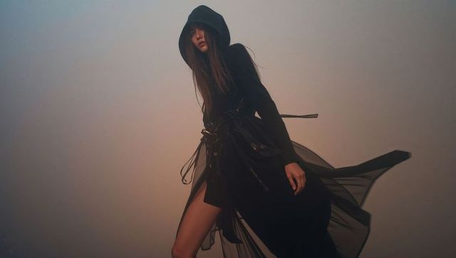 Asian model wearing black hooded coat with sheer panels posing in foggy fashion scene