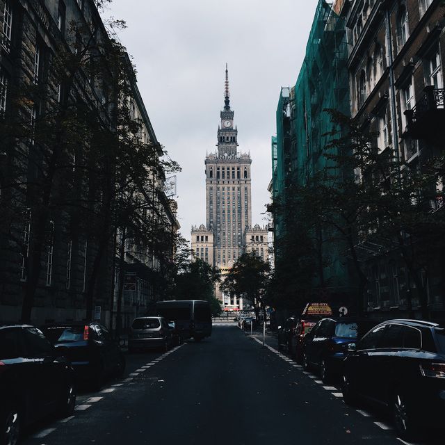 Photo shows a city street filled with parked cars leading up to a tall historical building. Surrounding buildings exemplify varied architectural styles. Overcast sky adds a moody atmosphere. Ideal for use in urban studies, architectural analysis, travel brochures, and educational materials.