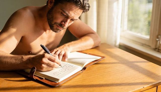 Shirtless man writing in leather journal at sunlit wooden desk