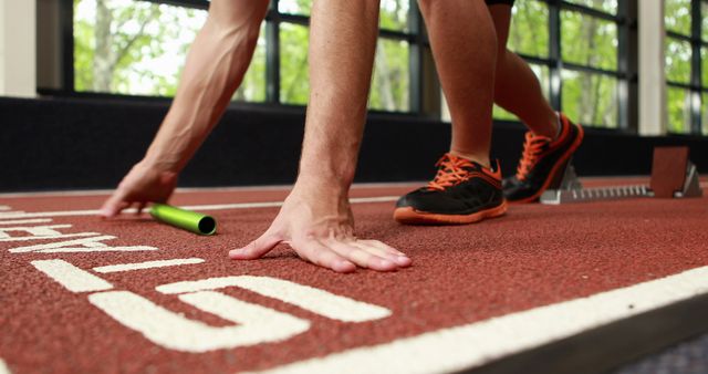 Runner kneeling at starting line with baton, getting ready to start a relay race on a track. Useful for topics on sports, athletics, preparation, and teamwork. Can be used by sports magazines, motivational posters, and fitness blogs.