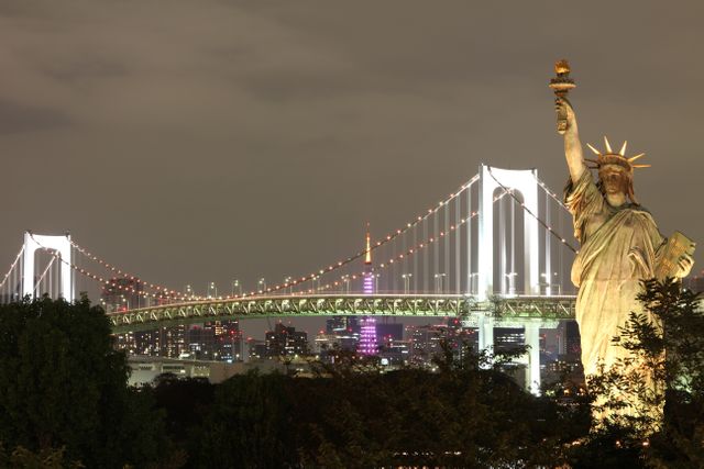 Statue of Liberty replica standing illuminated against nighttime backdrop of Rainbow Bridge and Tokyo skyline. Ideal for travel-related content, articles about Tokyo landmarks, posters promoting nighttime tourism.