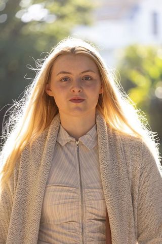 Young woman standing in sunlit park with backlit hair, cozy knit cardigan, soft bokeh