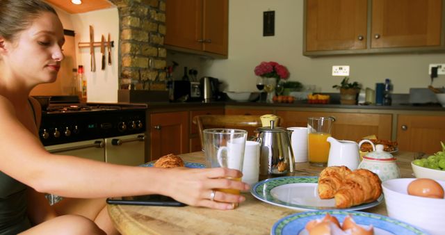A young Caucasian woman is seated at a kitchen counter with a variety of breakfast items, including croissants and orange juice, with copy space. Her relaxed posture and the homey setting suggest a leisurely morning meal.