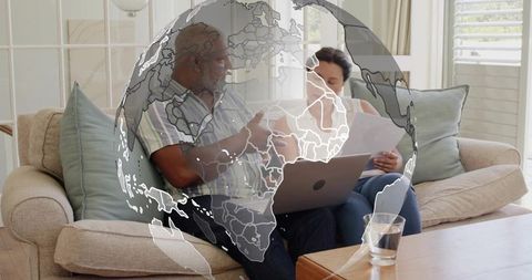 Mature couple reviewing documents on laptop on sofa with globe overlay for global collaboration