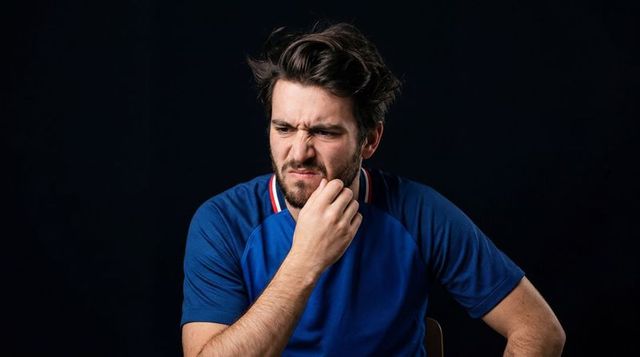 Young man grimacing and holding chin in blue polo, pensive moody headshot for editorial