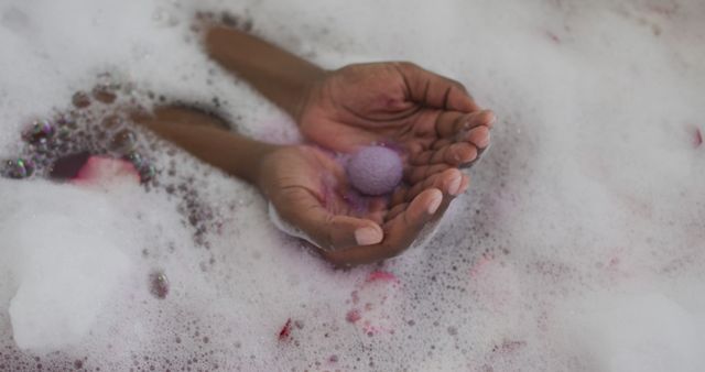 Hands are gently holding a purple bath bomb in a foamy, rose petal-filled bath. The scene conveys relaxation, self-care, and tranquility, making it ideal for wellness, spa, and skincare themes. This can be used in marketing materials for bath products, spa treatments, and health and wellness promotions.