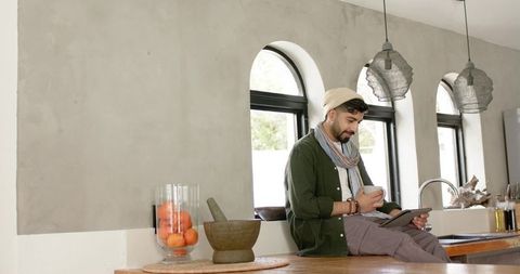 Indian man sitting on kitchen island using tablet holding coffee in minimalist daylight interior