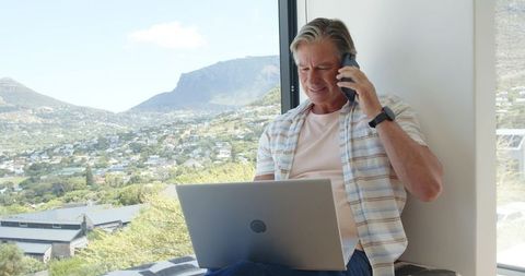 Mature man working remotely on laptop and phone from window seat with scenic valley view