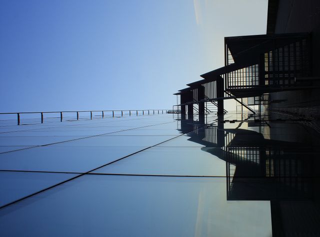 Displaying the sleek, reflective glass facade of a modern city building, this image captures the intersection of urban architecture and minimalist design. The reflection of the sky on the building's surface adds a serene touch and highlights the futuristic aspects of city planning and development. This picture can be used to depict themes such as modern architecture, urbanization, or real estate development.