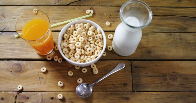 This image can be used in contexts related to food and healthy eating, such as promoting nutritious breakfast choices, dietary guides, or lifestyle blogs. It offers a neat arrangement of a bowl of cereal, a glass of orange juice with a straw, a small bottle of milk, and a spoon, all placed on a rustic wooden table. Ideal for food-related websites, health and wellness content, and meal planning illustrations.