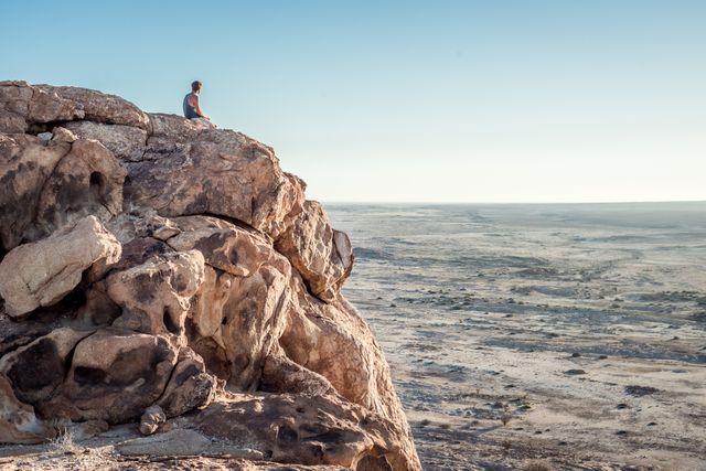 Individual sitting on edge of high rocky cliff, staring into vast desert landscape with clear sky and horizon. Ideal for travel advertisements, meditation guides, wilderness exploration features, or illustrating themes of solitude and reflection.