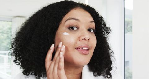 Woman applying facial moisturizer with fingertips at bathroom window for glowing skin