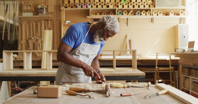 African american male carpenter wearing an apron carving wood with chisel at carpentry shop. carpentry, craftsmanship and handwork concept