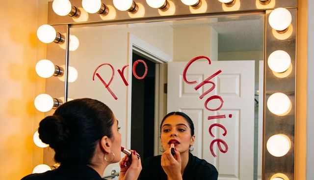 Woman applying red lipstick at lighted vanity mirror with pro choice message