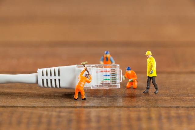 Miniature engineers and workers are seen repairing a LAN cable on a wooden table. This image can be used to represent concepts related to technology, internet maintenance, network repair, and teamwork. It is ideal for illustrating articles, blog posts, or advertisements about IT services, network solutions, or technical support.