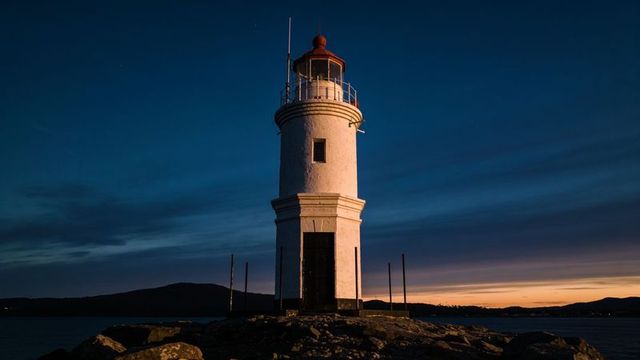 Standing lighthouse tower on rocky islet at dusk with deep blue twilight sky and sunset glow