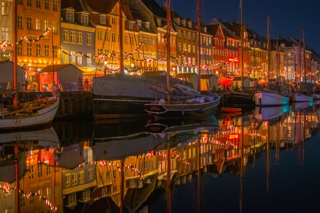 Colorful boats docked along the Nyhavn canal in Copenhagen, brightly illuminated waterfront houses reflecting in water during nighttime. Useful for travel blogs, tourism promotions, cityscape photography, and European destination advertisements.
