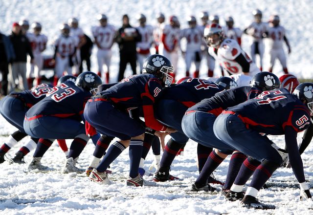 Image shows high school football teams playing a game in the snow. Players are seen lined up, ready for action, showcasing teamwork and competitive spirit. Ideal for use in articles, blogs, and campaigns focusing on youth sports, winter sports activities, school spirit, teamwork, and athletic determination.