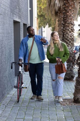 Couple walking with bicycle on urban sidewalk while man pointing and woman carrying bag